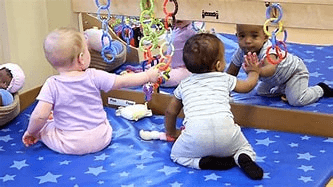 Babies playing on a blue mat with toys near a mirror.